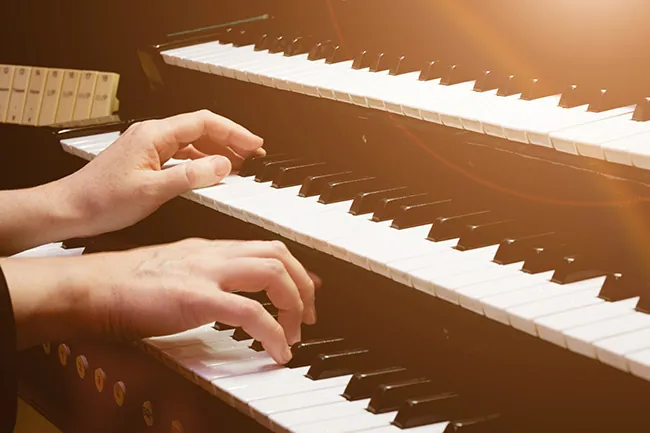 Close-up of hands playing an organ, focusing on the black and white keys. The image is illuminated with a warm, soft light, creating a serene atmosphere.