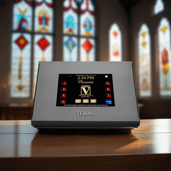 A Concerto Carillon music system displayed on a wooden table inside a church, with colorful stained glass windows in the background. The device features a touchscreen interface showing the brand logo and various music options.