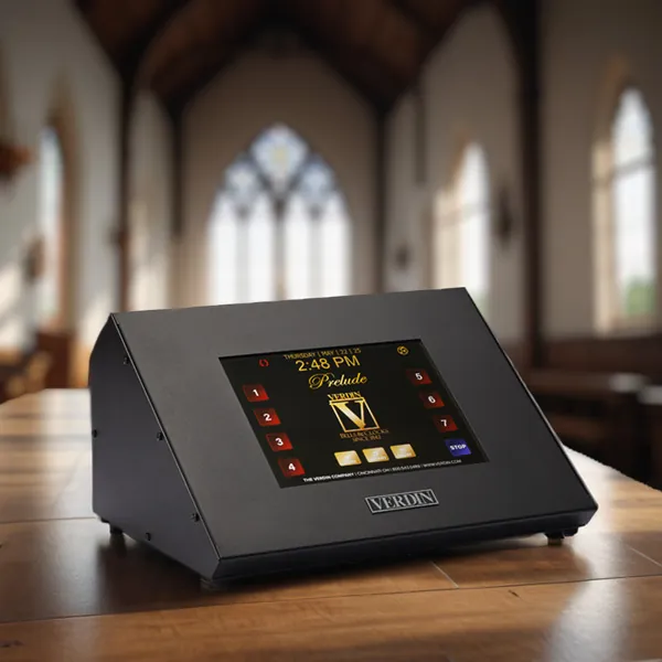 A Prelude Carillon device displayed on a wooden table inside a spacious, well-lit church. The device features a digital screen with colorful buttons and a clock, set against a backdrop of tall, arched windows and wooden pews.