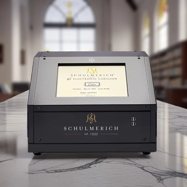 A Schulmerich Symphony carillon device on a white and grey marble table. In the background, white walls and large windows brighten the room. The console has a digital display, indicating its functionality for playing carillon music.